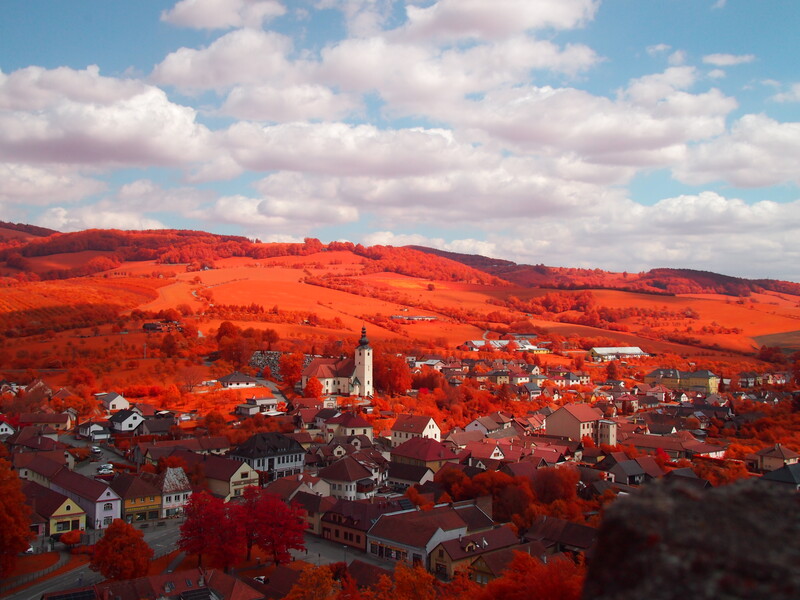 A village surrounded by hills and fields. The grass and trees are bright red, the sky is a light blue.